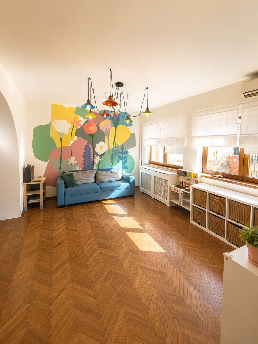 Sala de joacă a centrului: mural cu flori pictat pe perete, canapea albastră, parchet și cutii cu jucării